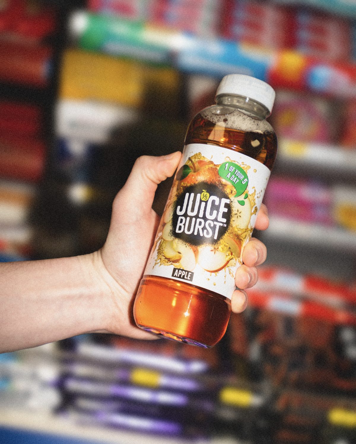 A hand holding a bottle of Juice Burst Apple in a corner shop aisle